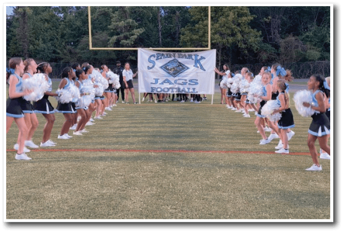 Two lines of cheerleaders in uniform with pom-poms face each other on a football field with a banner reading “Spirit Bank Jags Football" in the background. Description generated by AI