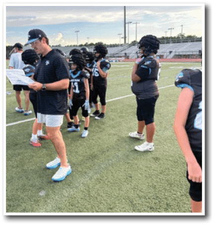 A group of people wearing football helmets and uniforms stand on a sports field with one adult in a black shirt and shorts holding a paper. Description generated by AI