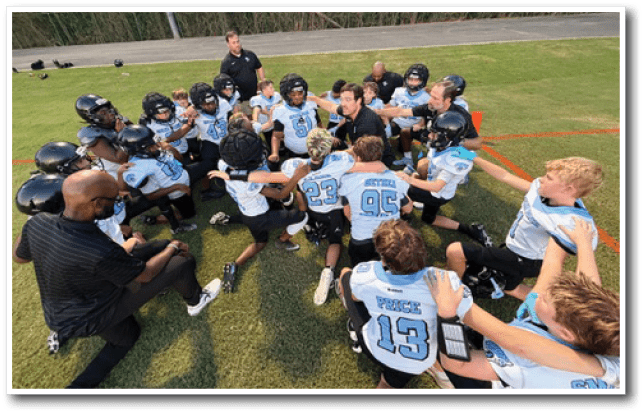 A group of children in football uniforms and helmets are gathered in a circle on a grassy field with adults, appearing to be in a team huddle or discussion. Description generated by AI