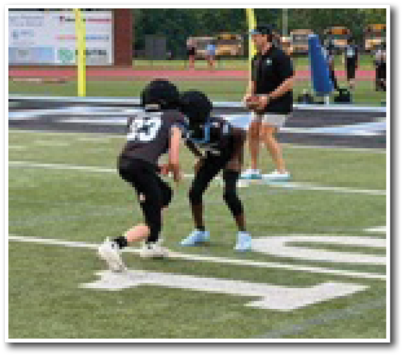 Two people in football gear are facing each other on a field near the 10-yard line while others and a coach stand in the background near the goalpost. Description generated by AI