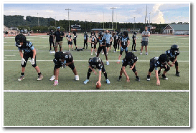 A group of people in football uniforms are lined up on a field in a ready position with coaches and other players standing behind them. Description generated by AI