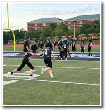 Two people in black sports uniforms are running on a football field with others and a coach standing near the end zone and school buses in the background. Description generated by AI