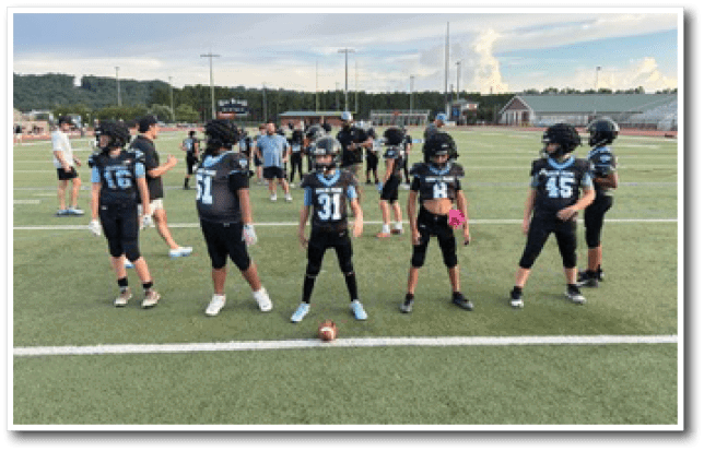 Children in football uniforms and helmets stand on a field in a line with a football placed on the ground in front of them. Description generated by AI
