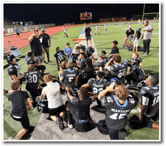 A group of people in sports uniforms are gathered on a field, listening to two standing adults who appear to be coaches giving instructions. Description generated by AI