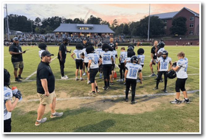 Children in football uniforms and adults stand on a grassy field near a stadium with spectators during sunset. Description generated by AI