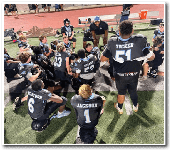A group of people in black football uniforms with white numbers are gathered on a field, some sitting and some standing, while a person in a cap appears to be speaking to them. Description generated by AI
