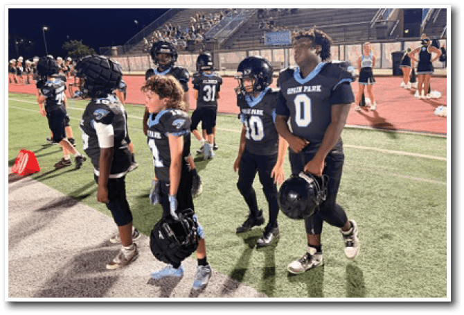 Children in football uniforms stand on a field at night near the sidelines with a stadium and spectators in the background. Description generated by AI