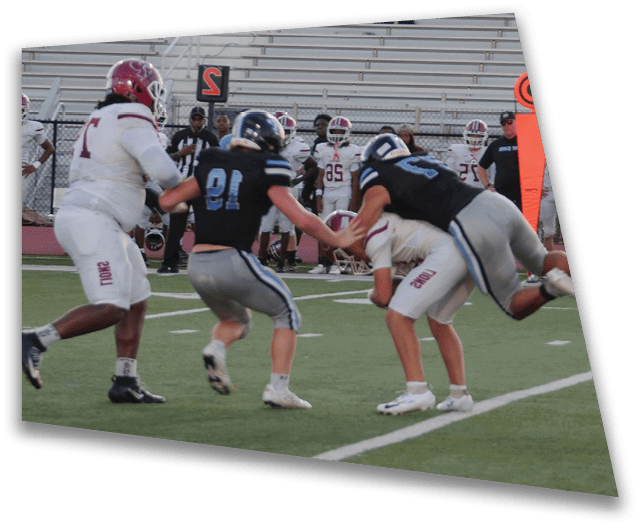 Two football players in black uniforms tackle a player in a white uniform on a field with empty bleachers and standing people in the background. Description generated by AI