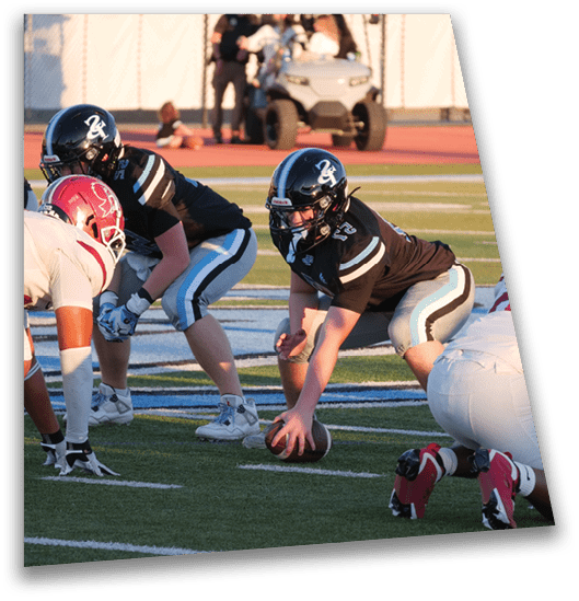 A person in a black football uniform is crouched on a field holding a football while another person in a white uniform with red helmet is positioned nearby in a defensive stance. Description generated by AI