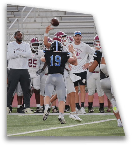 A group of people in football uniforms stand on a field with one person in a black jersey raising an arm while others in white jerseys and a person in casual sportswear watch. Description generated by AI