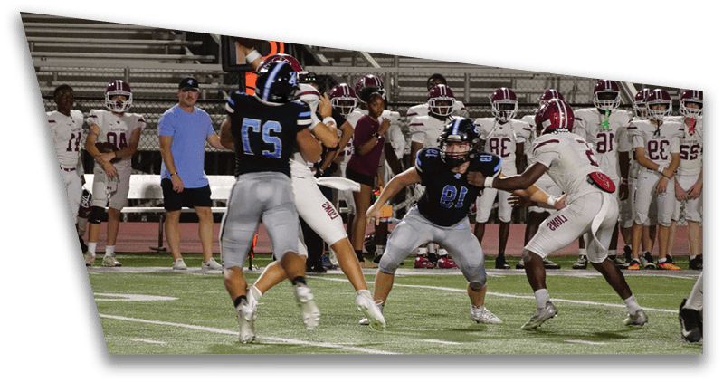 People in football uniforms are actively playing on a field with others standing on the sidelines watching. Description generated by AI