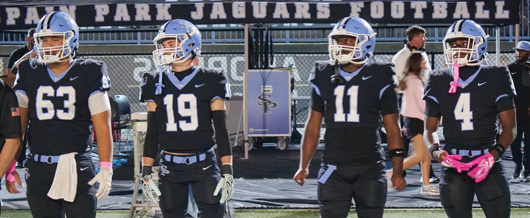 Four people in black football uniforms with helmets stand side by side on a field with a scoreboard and spectators in the background. Description generated by AI
