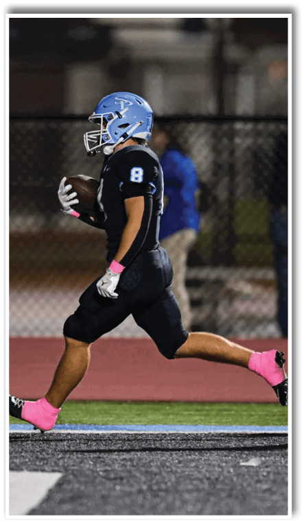 A person wearing a blue helmet and black football uniform with pink socks and gloves is running on a football field while holding a football. Description generated by AI