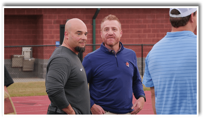 Two adults stand on a track field engaged in conversation, one wearing a dark long-sleeve shirt and the other in a blue pullover with a patterned collar. Description generated by AI