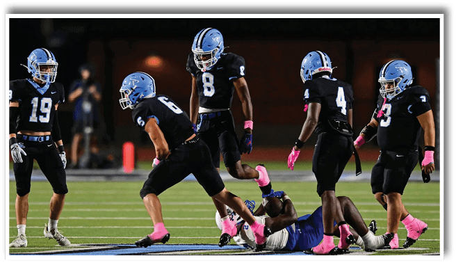 Five people in black sports uniforms with blue helmets and pink gloves are on a football field, with one person lying on the ground and the others standing or moving around them. Description generated by AI