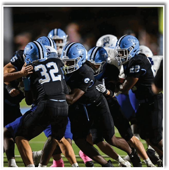 Several football players wearing black uniforms and light blue helmets are engaged in a play on a field under stadium lights. Description generated by AI