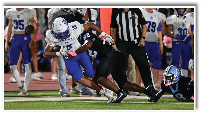 A person in a white and blue football uniform is running with the ball while being tackled by a person in a black uniform on a football field with other players and a referee in the background. Description generated by AI