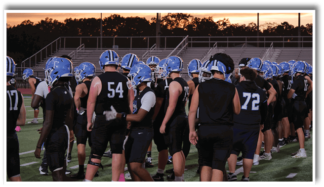 A group of people wearing blue helmets and black sports uniforms are gathered on a field at sunset with empty bleachers in the background. Description generated by AI