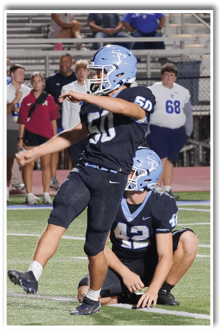 Two people in football uniforms and helmets are on a field with one holding the ball and the other kneeling to assist. Description generated by AI