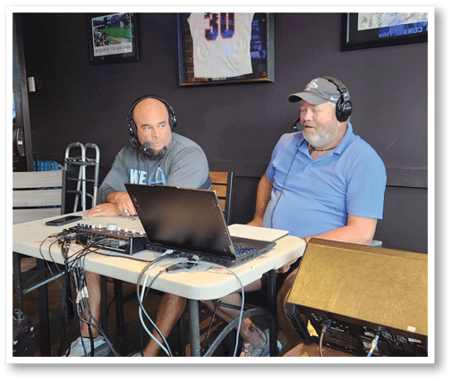 Two adults wearing headsets are sitting at a table with a laptop and audio equipment, engaged in conversation or broadcasting. Description generated by AI