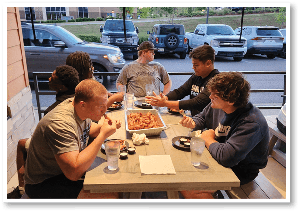 Four people sit around a table eating chicken wings from a large aluminum tray with dipping sauces and drinks nearby. Description generated by AI