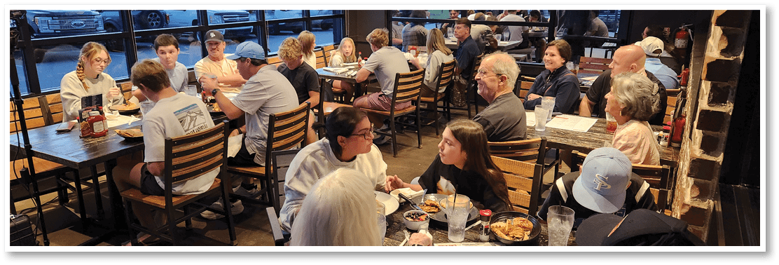 People are sitting and eating at wooden tables and chairs in a busy casual restaurant setting. Description generated by AI