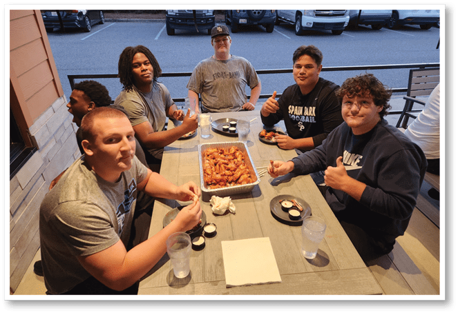 People sitting around a table eating chicken wings with dipping sauces and giving thumbs up. Description generated by AI