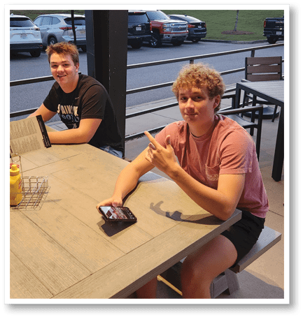Two people sit at a wooden table near a window, one wearing a black graphic t-shirt and the other wearing a reddish shirt while pointing with their hand. Description generated by AI