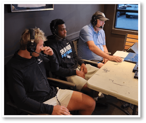 Three people wearing headsets are seated at a table with audio equipment, engaged in what appears to be a live broadcast or recording session. Description generated by AI