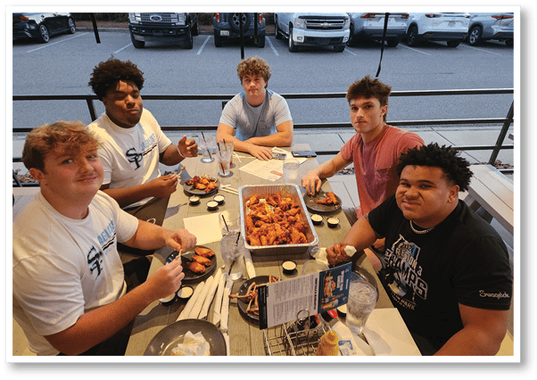 Five people sit around a table with a large tray of chicken wings and several dipping sauces, some holding wings and drinks. Description generated by AI