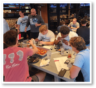 A group of people are gathered around a table with food, some standing and some sitting, engaging in conversation and eating in a casual indoor setting. Description generated by AI