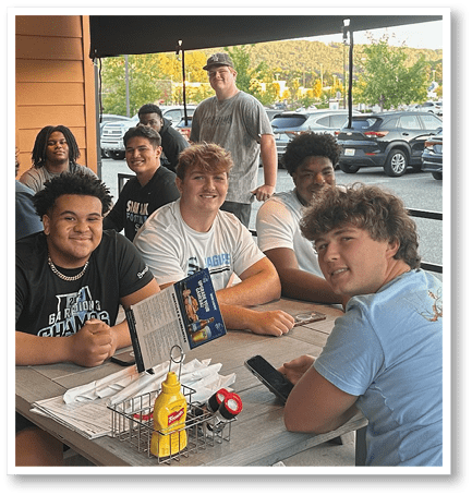 A group of people with casual clothing are sitting and standing around a table outdoors near a parking lot, smiling and engaging with each other. Description generated by AI
