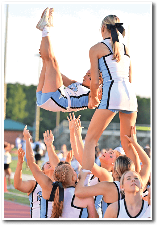 A group of people in cheerleading uniforms are performing a stunt outdoors on a sports field with one person being lifted and flipped in the air while others support and catch them. Description generated by AI