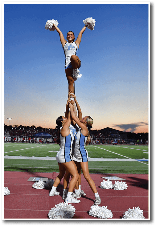 People in cheerleading uniforms perform stunts and hold pom-poms on the sidelines of a sports field during sunset. Description generated by AI