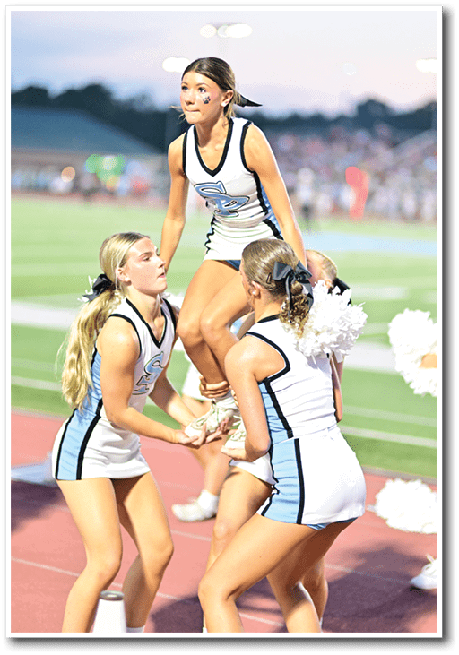 A person in a cheerleading uniform with a paw print face sticker is being lifted by two others on a sports field during an outdoor event. Description generated by AI