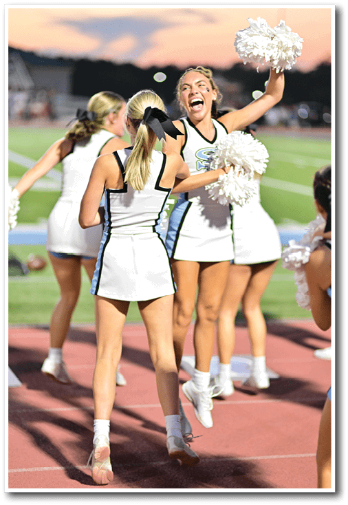 Three people in cheerleading uniforms are interacting outdoors with one person laughing joyfully and another with a black bow in their hair. Description generated by AI