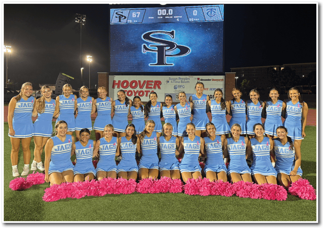 A group of adults in blue cheerleading uniforms with pink pom-poms pose on a football field in front of a scoreboard showing a final score at Spain Park High School. Description generated by AI