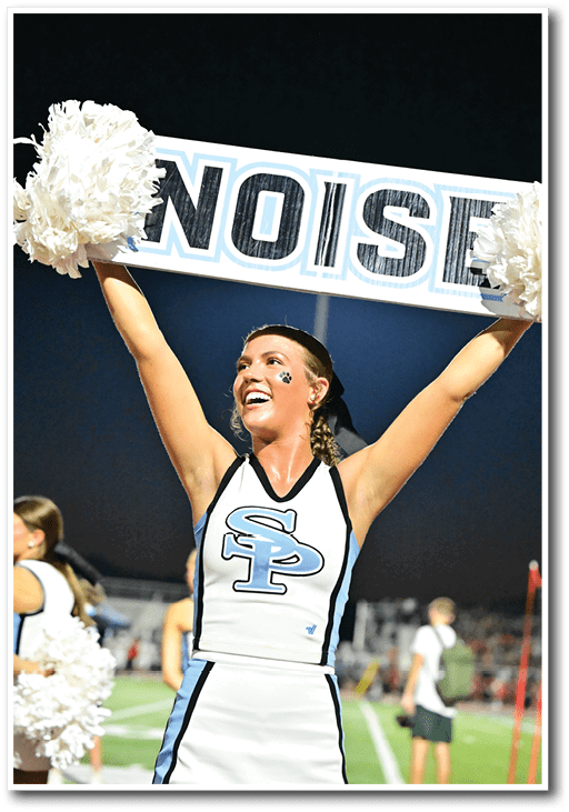 A person in a cheerleading uniform with “SP" on the front is smiling and holding up a sign that says "NOISE" with pom-poms on each side at a nighttime outdoor sports event. Description generated by AI