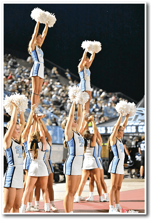 A group of cheerleaders in blue and white uniforms perform a coordinated stunt with some lifted high holding pom-poms at a nighttime sports event. Description generated by AI