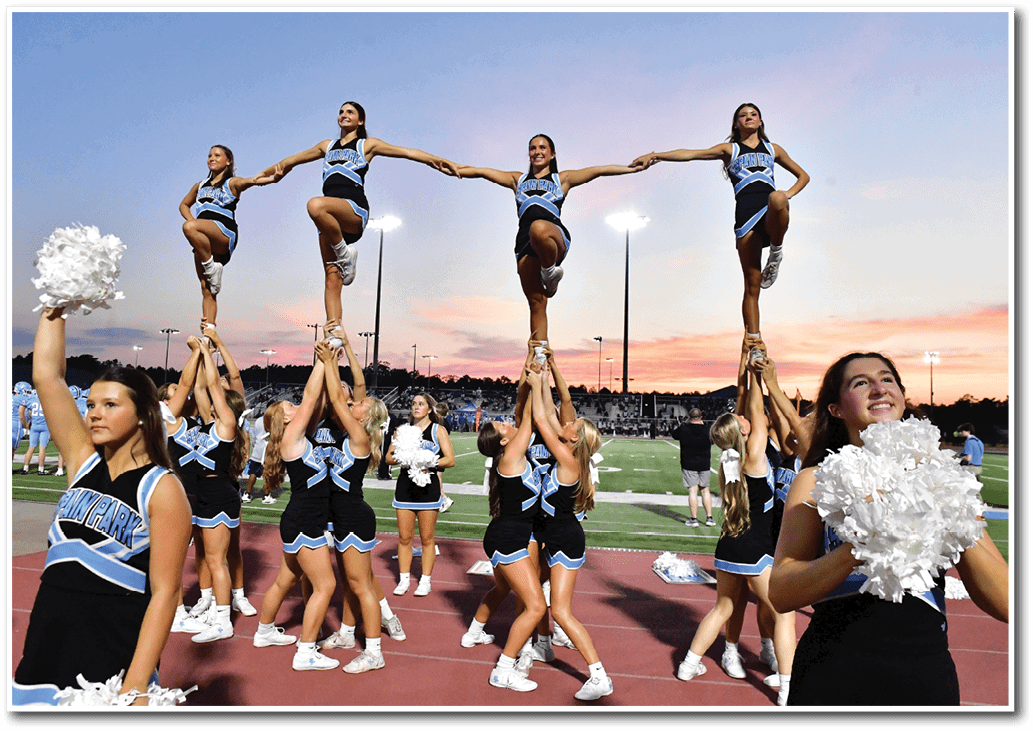 A group of cheerleaders in matching uniforms perform a coordinated stunt on a sports field at sunset with some holding pom-poms and others lifted in the air. Description generated by AI