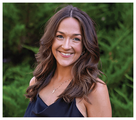 A smiling person with long wavy hair wearing a black sleeveless top and a necklace with a cross pendant stands outdoors in front of green foliage. Description generated by AI