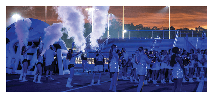 People dressed in sparkly outfits and boots are performing a dance routine on a field at night with a crowd watching in the background. Description generated by AI