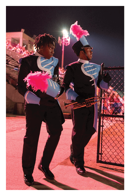 A person in a blue and black marching band uniform with a tall feathered hat is playing a saxophone on a football field at sunset. Description generated by AI