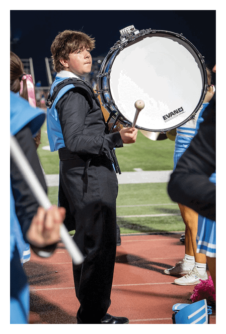 A person in a black marching band uniform is playing a large bass drum on a track field with other band members in blue uniforms nearby. Description generated by AI