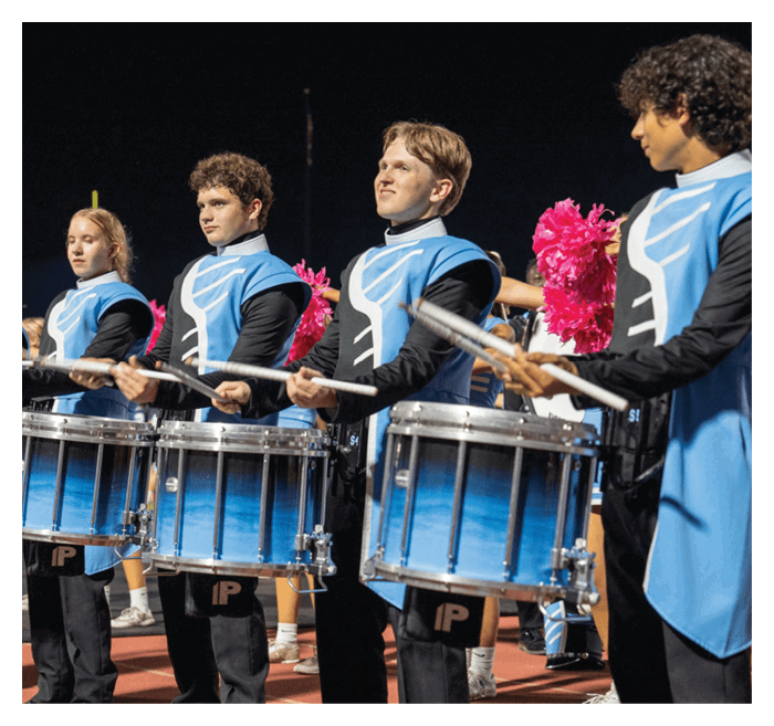 People wearing blue and black uniforms are playing snare drums in a line on a track field. Description generated by AI