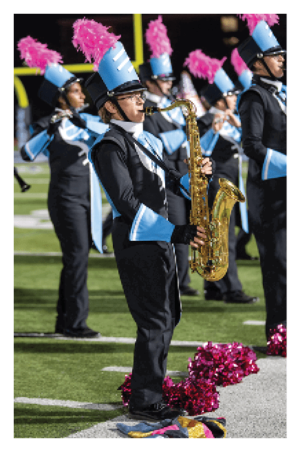 People in marching band uniforms with blue hats and pink plumes are playing instruments on a football field at night. Description generated by AI