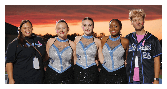 Five people stand side by side outdoors at sunset with three wearing sparkly blue and silver dance costumes and two wearing casual clothing. Description generated by AI