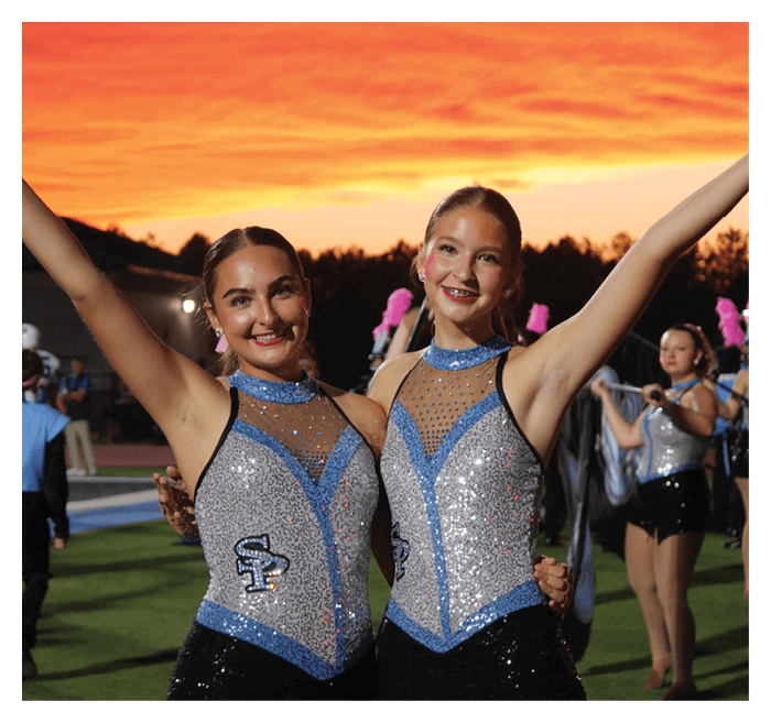 Two adults wearing sparkly blue and silver performance outfits smile with arms raised on a field at sunset while others in similar attire stand in the background. Description generated by AI