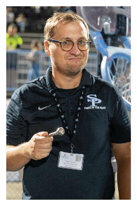 A person wearing glasses and a black polo shirt with a lanyard and badge is holding a small object and standing in front of a wheelchair. Description generated by AI