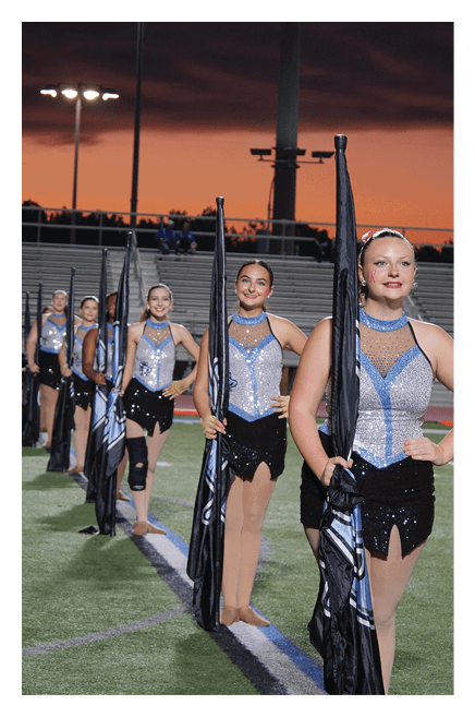 A line of people dressed in sparkly blue and black performance outfits stand on a sports field at sunset holding flags. Description generated by AI